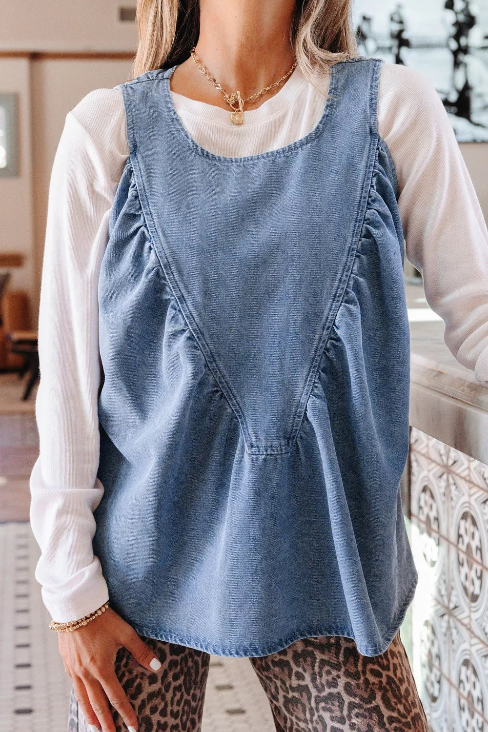 V-shaped pleated denim sleeveless top with a rounded neckline, styled over a white long-sleeve shirt.
