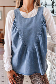 V-shaped pleated denim sleeveless top with a rounded neckline, styled over a white long-sleeve shirt.