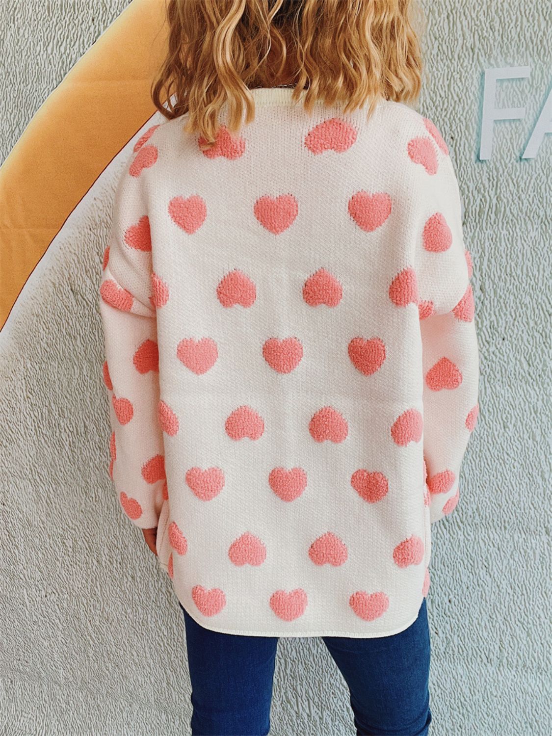 White acrylic cardigan with textured pink heart pattern, crew neck, long sleeves.