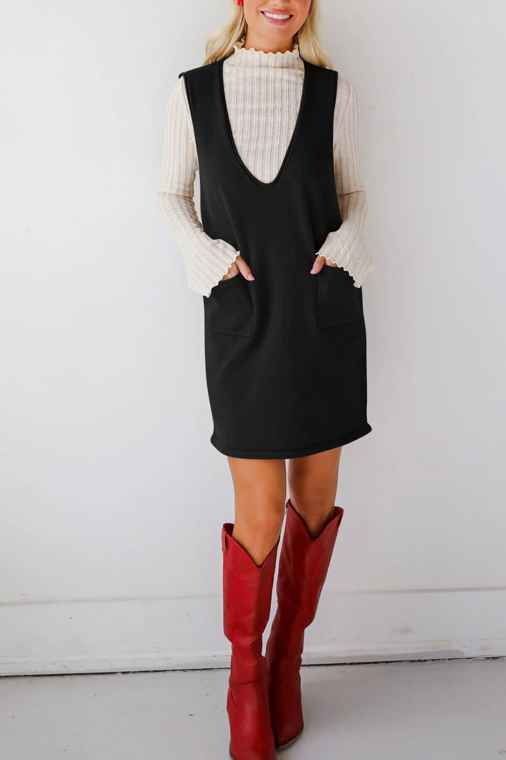 Black V-neck sweater dress with pockets, styled with a cream ribbed top and red boots.