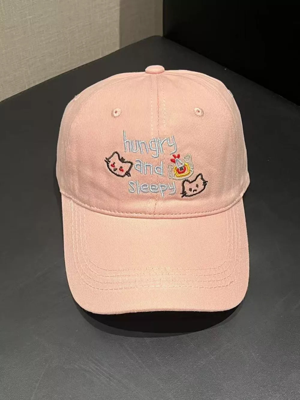 Light pink baseball cap with embroidered cats and text saying hungry and sleepy