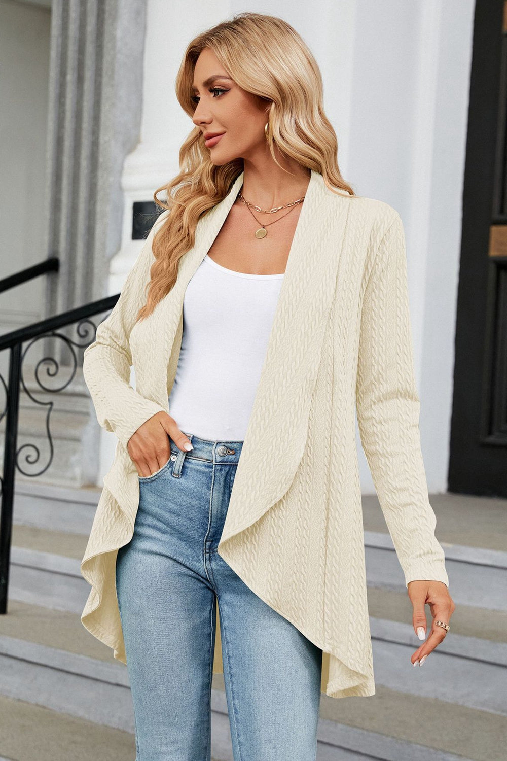 Cream cable knit drape-front cardigan with long sleeves, worn open over a white tank top and jeans.