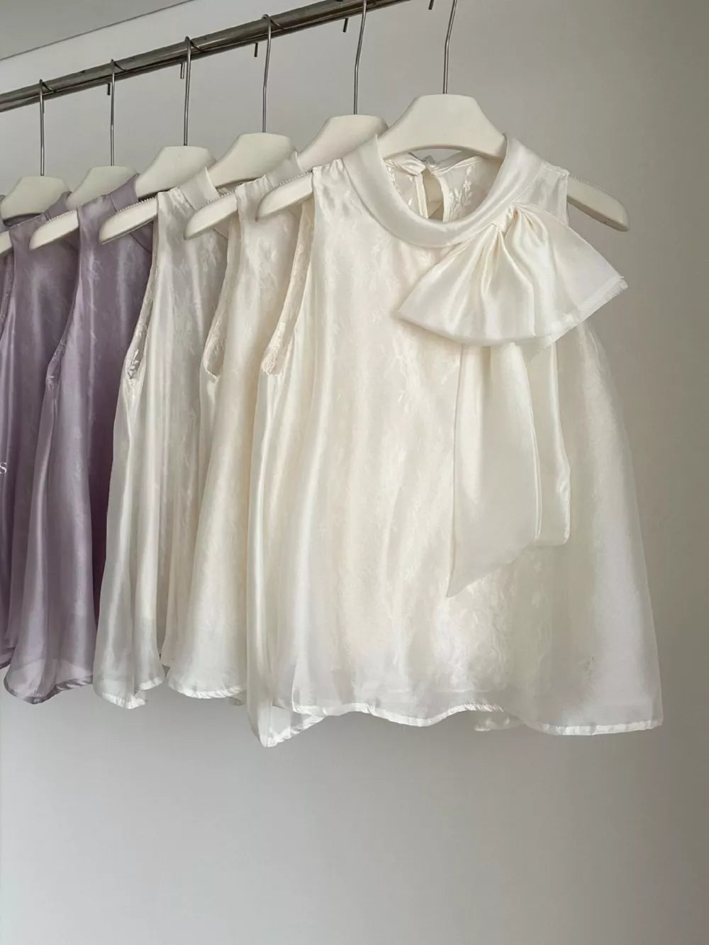 Sleeveless white shirt with a large bow detail and mock neck, available in purple and apricot.