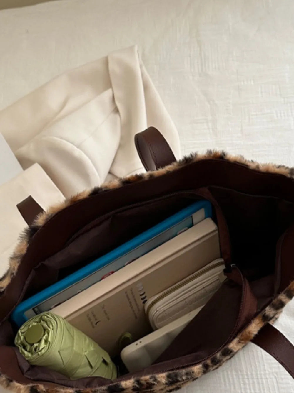 Fuzzy leopard print tote bag with brown handles, open and filled with a tablet, books, wallet, and umbrella.