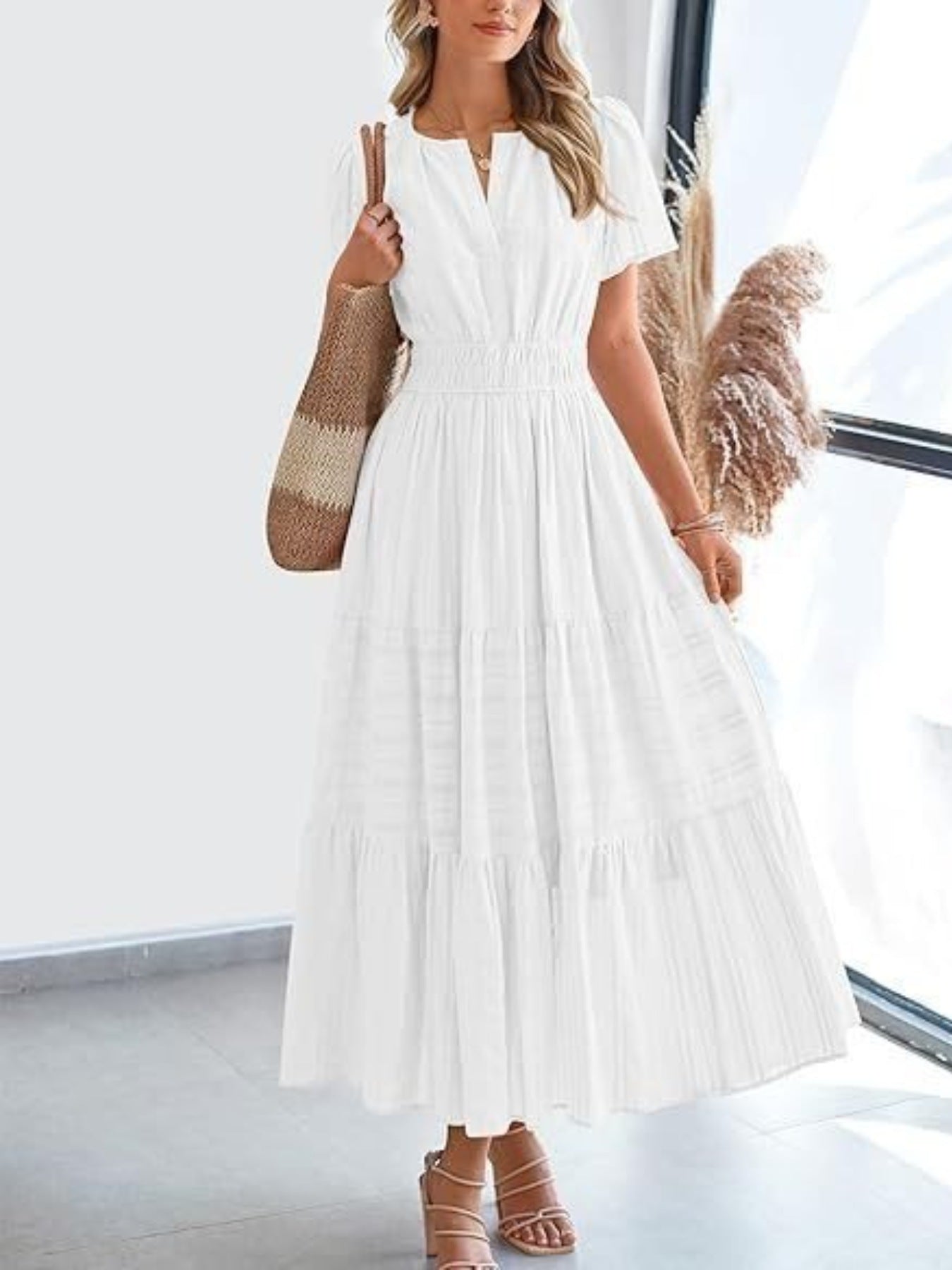White tiered V-neck maxi dress with smocked waist and short sleeves, styled with sandals and a tote bag.