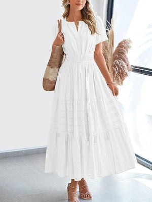White tiered V-neck maxi dress with smocked waist and short sleeves, styled with sandals and a tote bag.