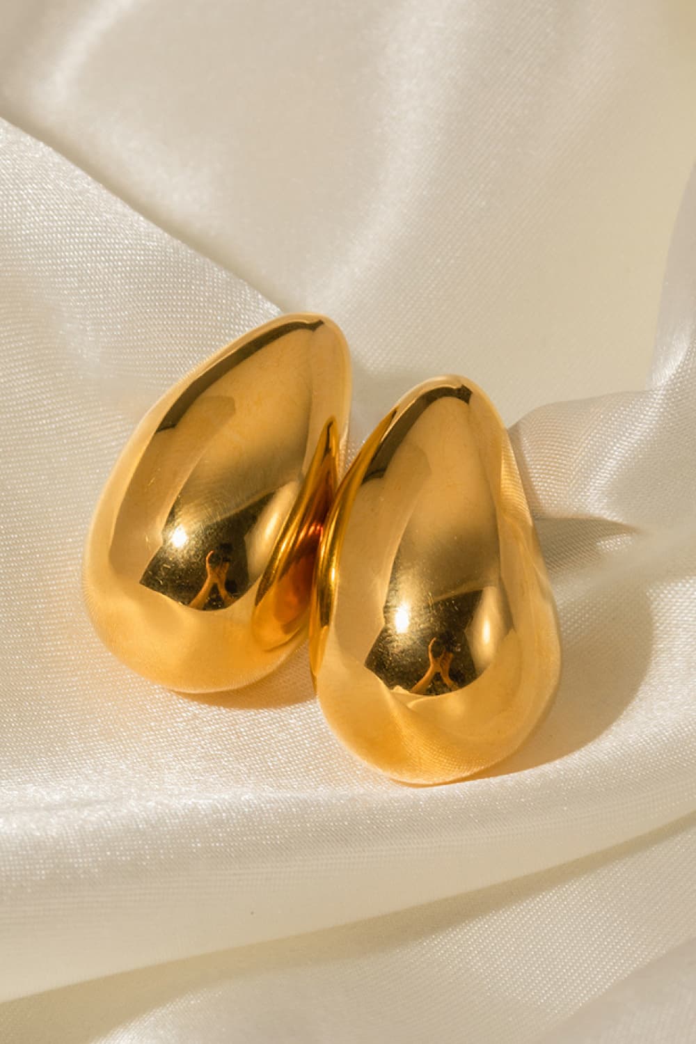 Sculptural gold teardrop stud earrings with a polished finish on satin fabric.