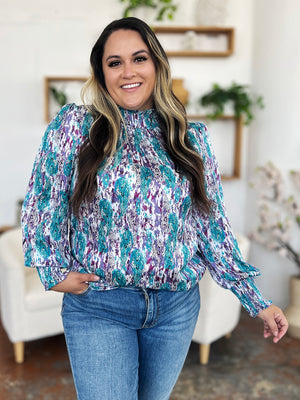 High neck abstract print blouse with smocked cuffs and hem in teal, purple, and white.