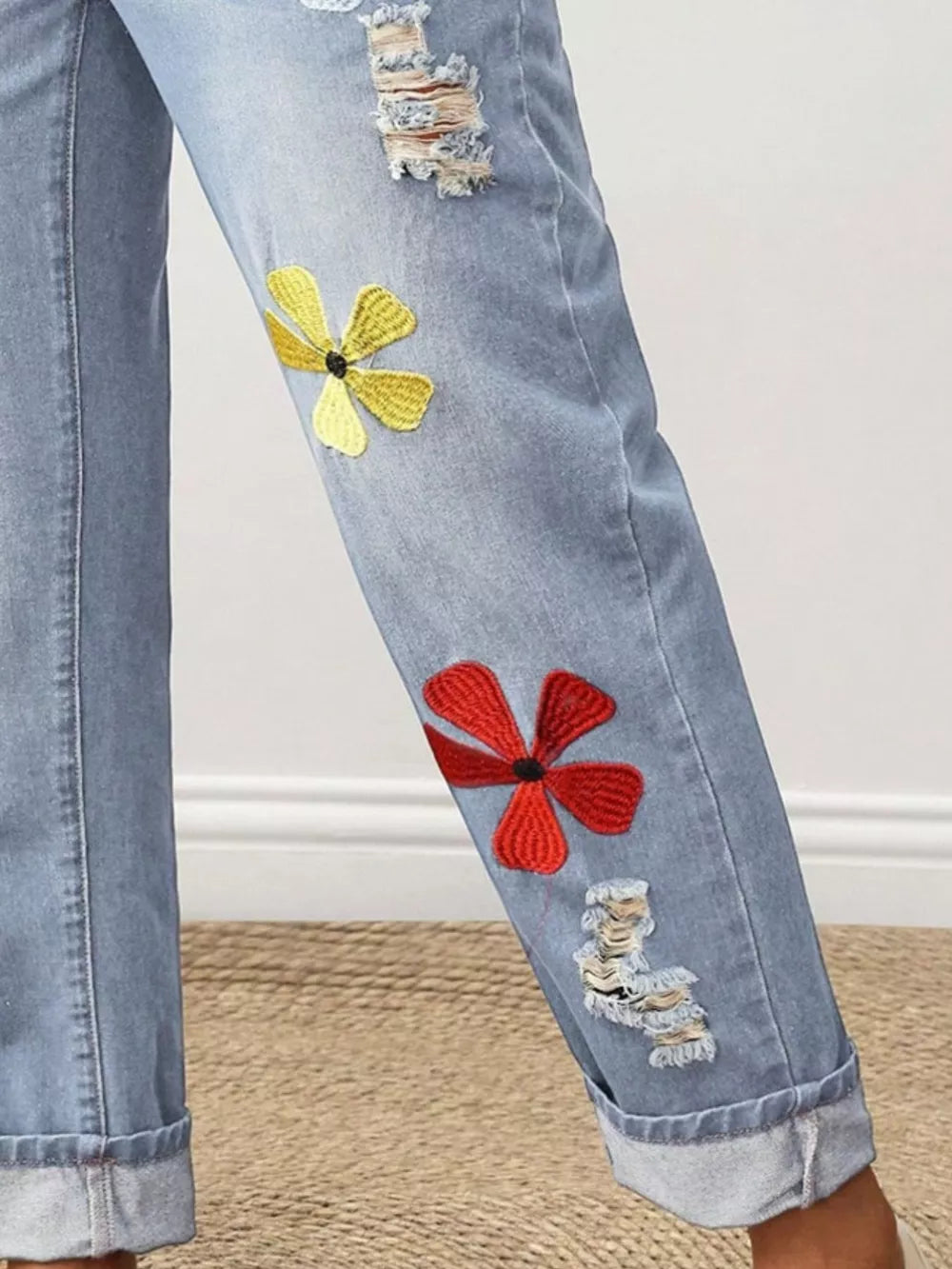 Light wash denim jeans with cuffed hems, distressed rips, and embroidered floral accents in yellow and red.