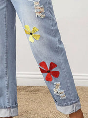 Light wash denim jeans with cuffed hems, distressed rips, and embroidered floral accents in yellow and red.