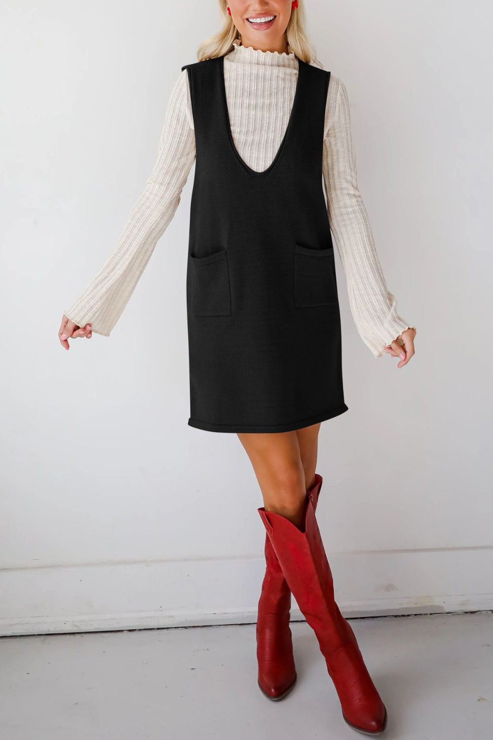 Black V-neck sweater dress with pockets, styled with a cream ribbed turtleneck and red boots.