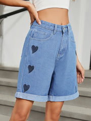Medium wash denim bermuda shorts with dark heart appliqués and cuffed hems.