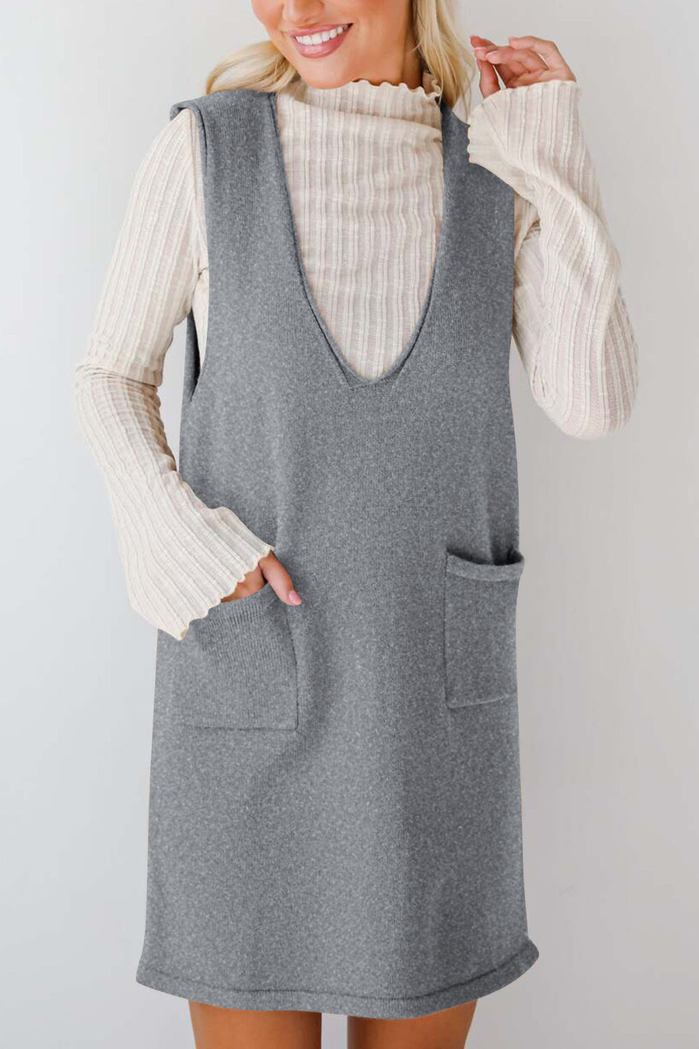 Sleeveless gray knit dress with V-neck and two front pockets, layered over a cream ribbed turtleneck.