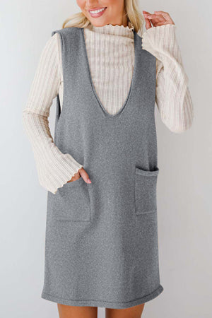 Sleeveless gray knit dress with V-neck and two front pockets, layered over a cream ribbed turtleneck.