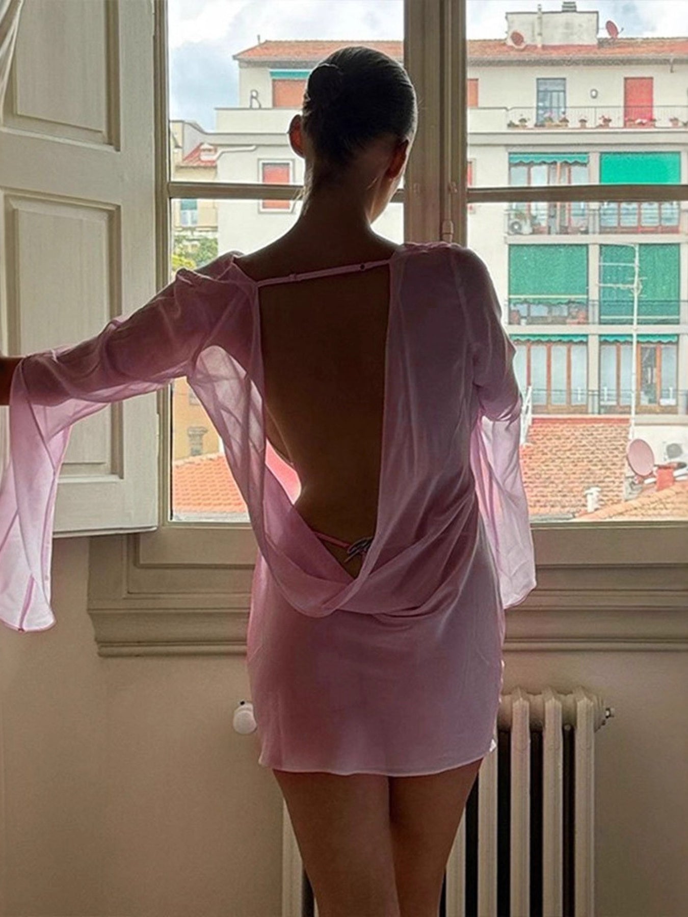 Pink chiffon backless mini dress with draped detail and long bell sleeves.