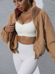 Tan fleece cropped funnel neck jacket with zipper detail, worn open over a white ruched sports bra.