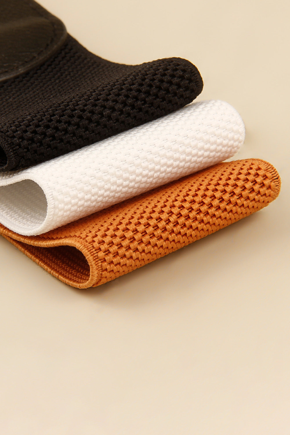 Black, white, and caramel elasticated belts with woven texture and sculpted buckle detail.