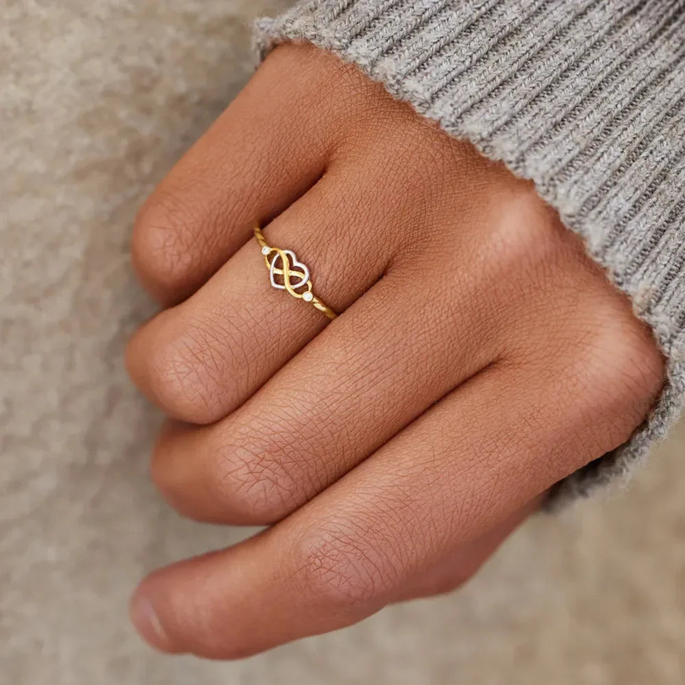Gold intertwined heart and infinity ring with small accent stones on a finger.