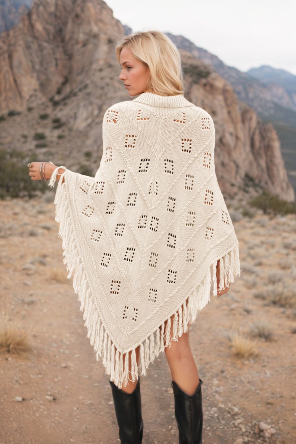 Cream knit poncho with geometric cutouts and fringe, acrylic material.