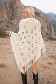 Cream knit poncho with geometric cutouts and fringe, acrylic material.