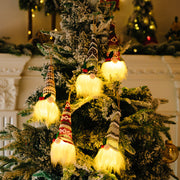 Illuminated gnome ornaments with knitted hats and fluffy white beards hang on a Christmas tree.