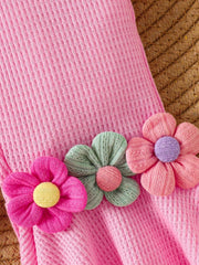 Pink cotton jumpsuit with smocked texture and floral appliques in pink, green, and pastel blue.