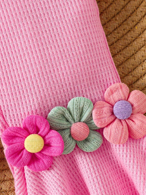 Pink cotton jumpsuit with smocked texture and floral appliques in pink, green, and pastel blue.
