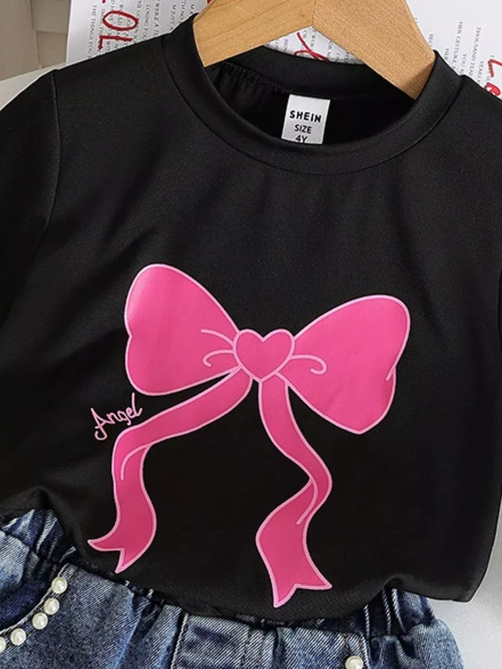 Black tee with pink bowknot graphic and "Angel" script, paired with denim shorts.
