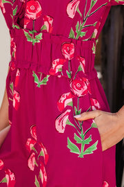 Charming floral tiered midi dress in magenta with red roses and green leaves, featuring a ruffled waist and pockets.