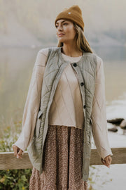 Sage quilted vest coat with diamond pattern and patch pockets, worn over a cream sweater and floral dress.