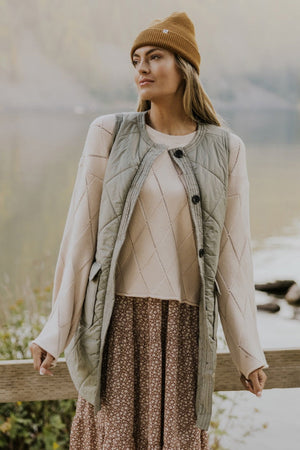 Sage quilted vest coat with diamond pattern and patch pockets, worn over a cream sweater and floral dress.