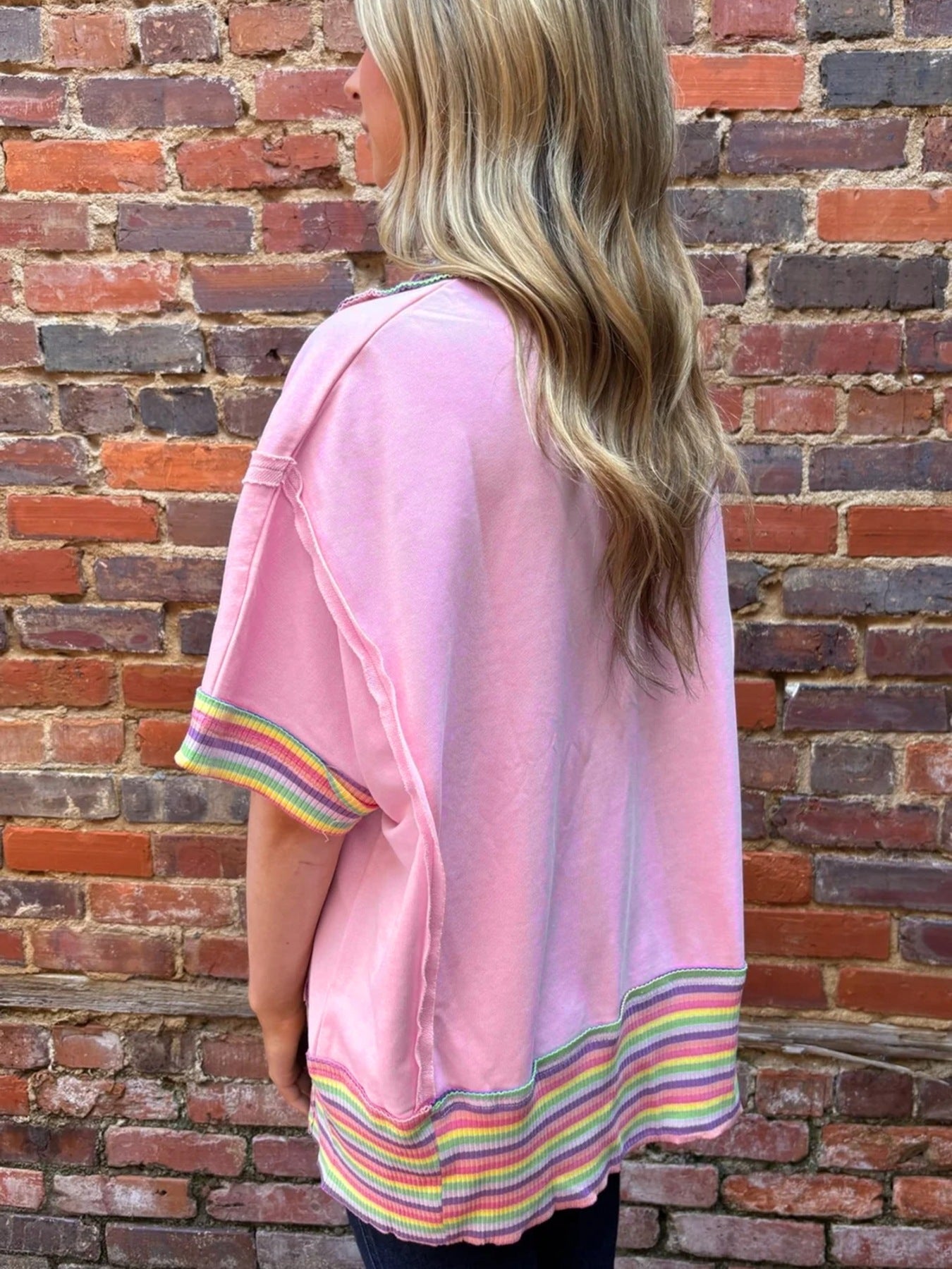 Pink cotton oversized V-neck top with rainbow striped cuffs and hem, exposed seams.