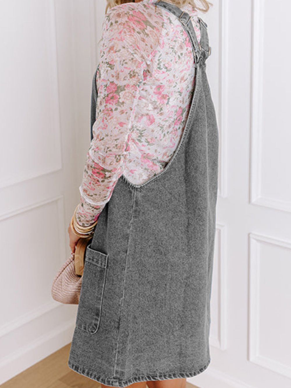 Faded gray denim pinafore dress with front pockets and adjustable straps, worn over a floral print top.