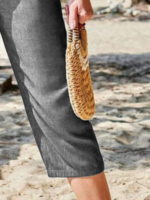 Dark gray denim capri trousers with elastic waist, shown with a straw clutch.