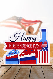 Patriotic wooden tabletop decor with "Happy Independence Day" text, cake slices, and a bottle with USA flag design.