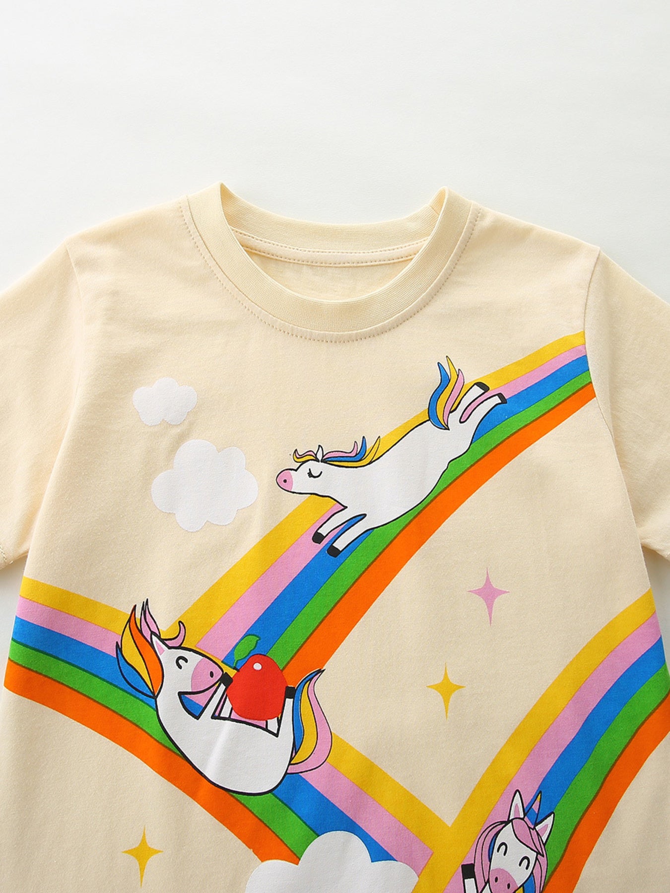 Cream cotton t-shirt with playful unicorns on a rainbow graphic, clouds, and stars.