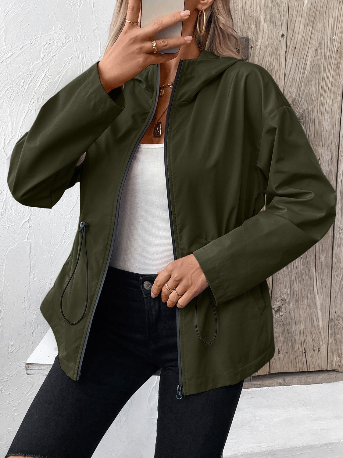 Army green utility jacket with drawstring waist and hood, zipped front, styled with a white tank top and black jeans.