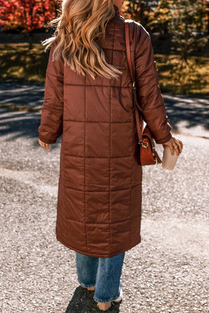 Brown quilted longline puffer coat with patch pockets, worn casually with jeans.