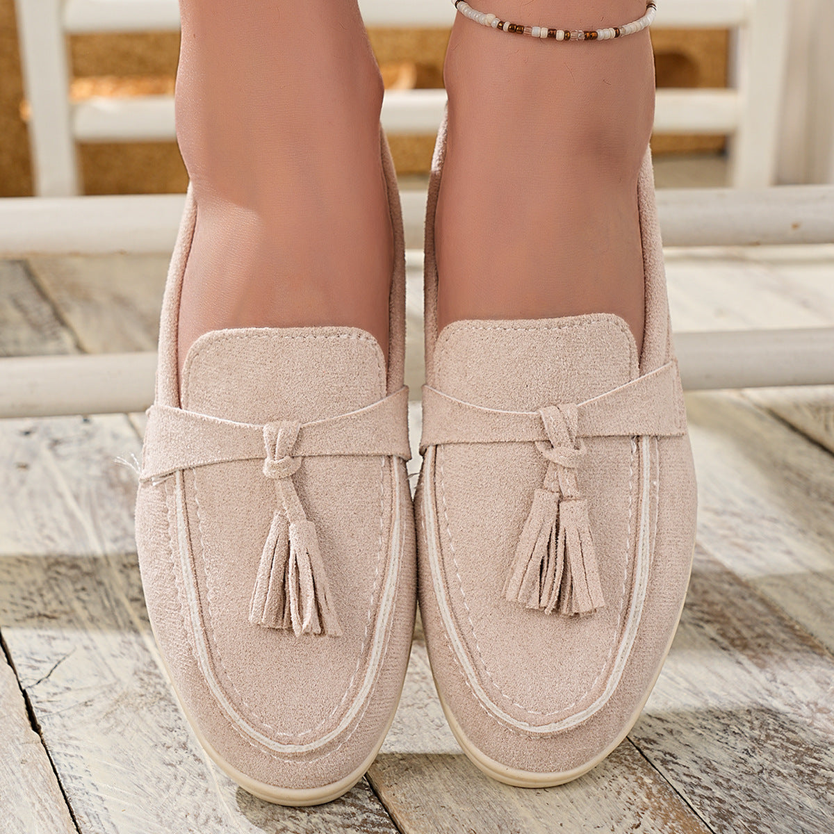 Beige faux-suede slip-on loafers with decorative tassels and contrast stitching.
