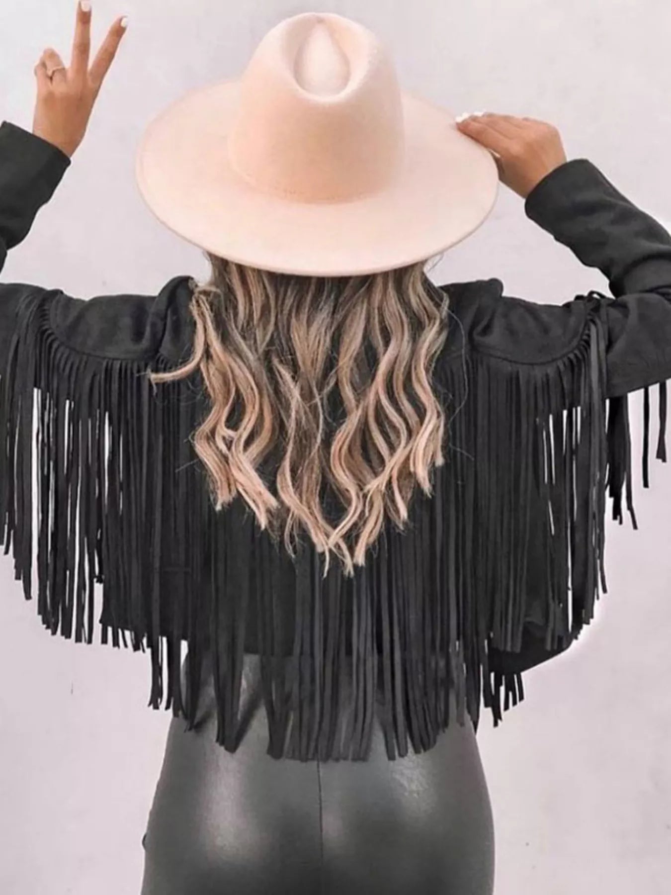 Black cropped jacket with fringe detail and open front, styled with a wide-brim hat.