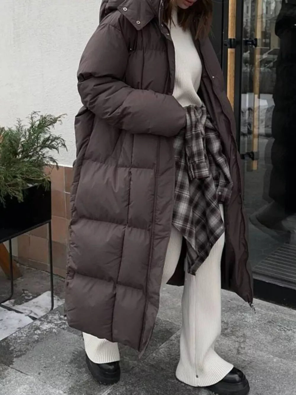 Dark brown long puffer coat with tie-belt detail and hood, worn open over a cream sweater and plaid shirt.