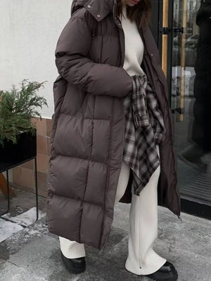 Dark brown long puffer coat with tie-belt detail and hood, worn open over a cream sweater and plaid shirt.