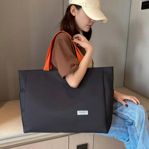 Spacious black canvas shoulder bag with bold orange straps and a small white fashion logo patch.