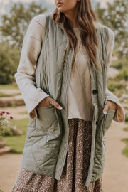 Sage quilted vest coat with patch pockets and button closure, styled over a cream sweater and floral dress.