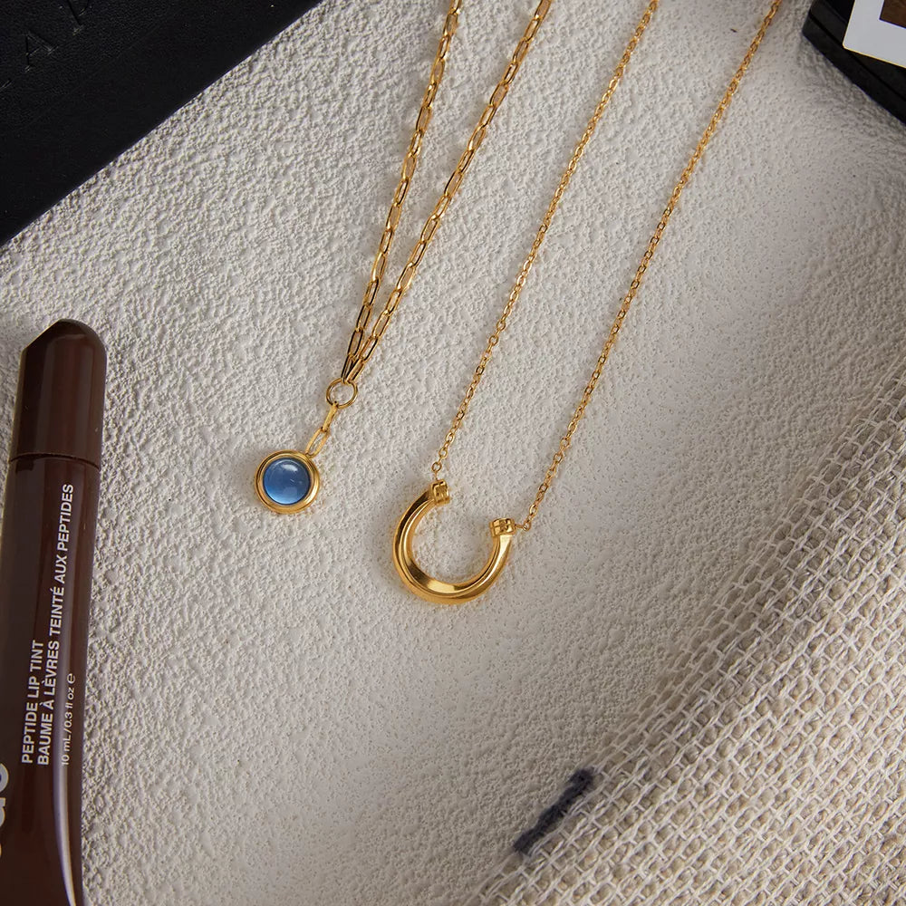 Elegant gold-plated layered necklaces, one with a blue stone pendant, the other a horseshoe charm.