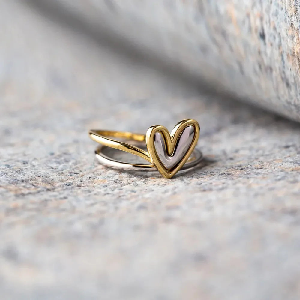 Two-tone heart outline ring in gold and silver, stacked on a stone surface.