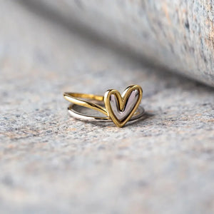 Two-tone heart outline ring in gold and silver, stacked on a stone surface.