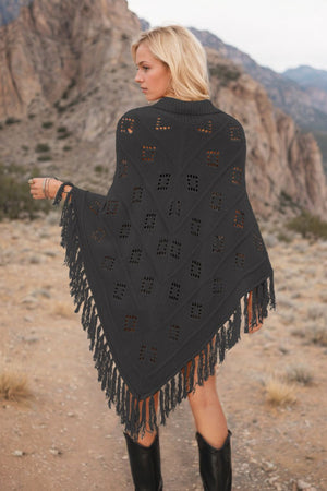 Black knit poncho with geometric cutouts and fringe, acrylic material.