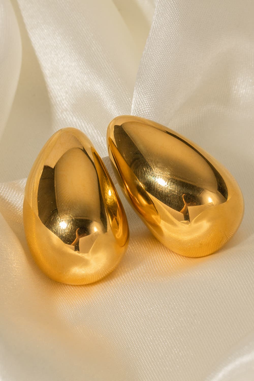 Gold teardrop stud earrings with a smooth, sculptural finish on satin fabric.