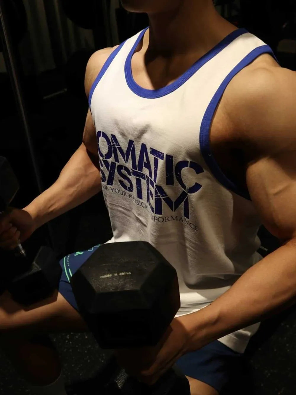 White tank top with blue trim and "SOMATIC SYSTEM" graphic, worn by a person lifting weights.